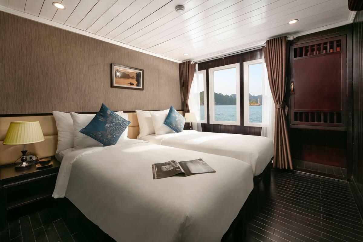 A hotel room with two beds, a nightstand with a lamp, a window with curtains, and a view of the water.