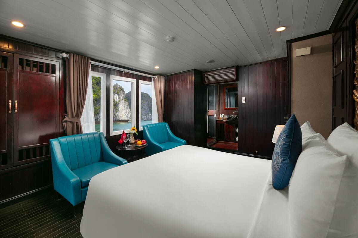 A hotel room with a large bed, two blue armchairs, a small table with fruit, and a view of the outdoors through large windows.