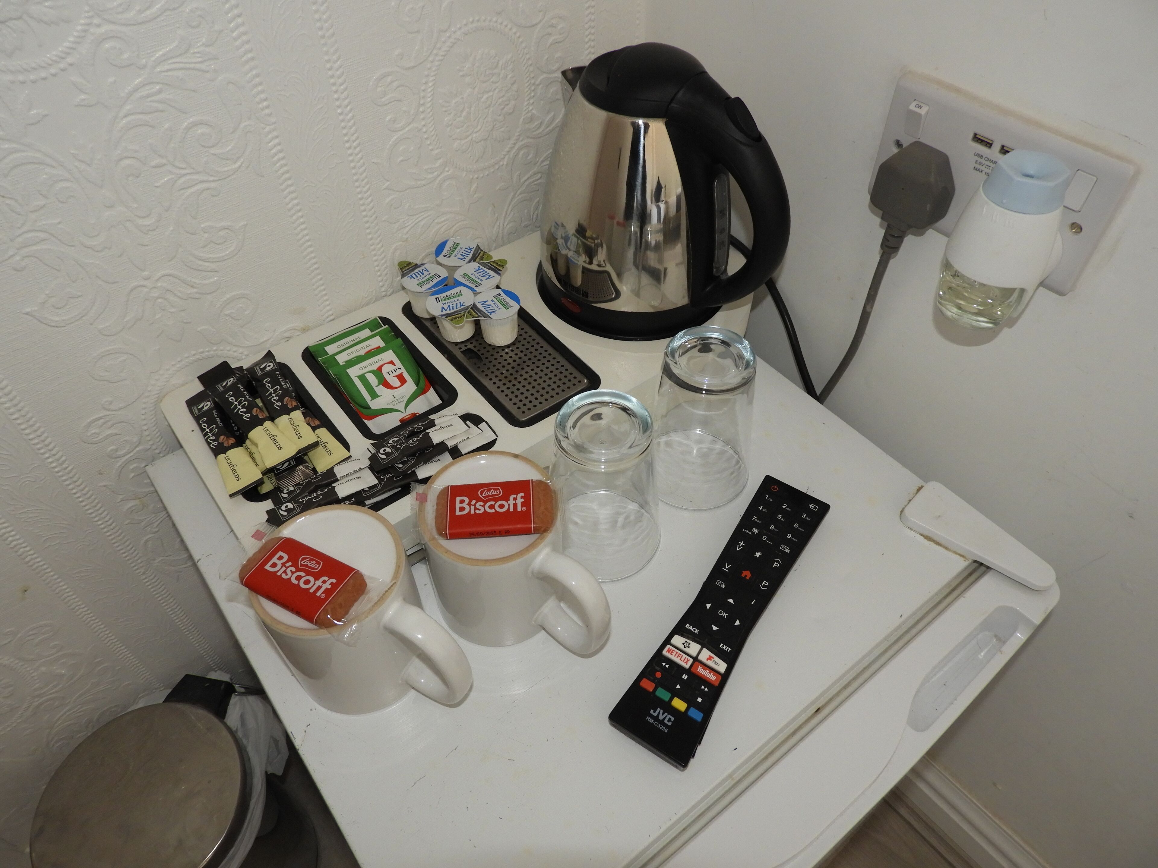 A small table with a coffee maker, two mugs, a remote control, and some food items.
