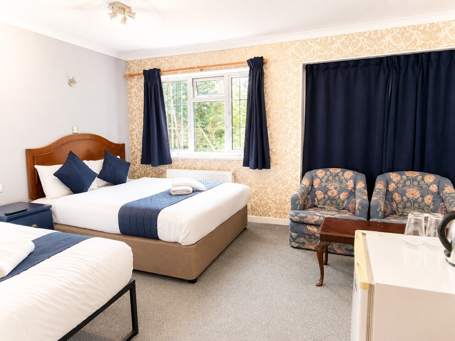 Quarto de hotel com cama, duas mesas de cabeceira, uma cadeira, uma mesinha e janela com cortinas.