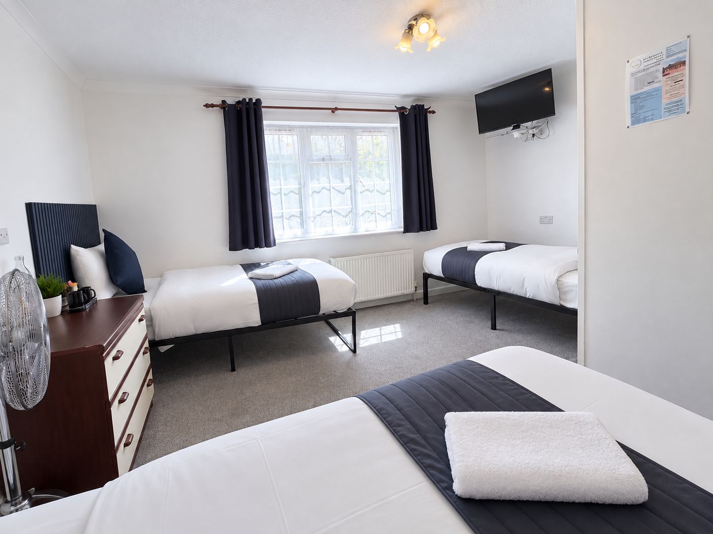 A hotel room with two beds, a dresser, a radiator, a TV, and a window with curtains.