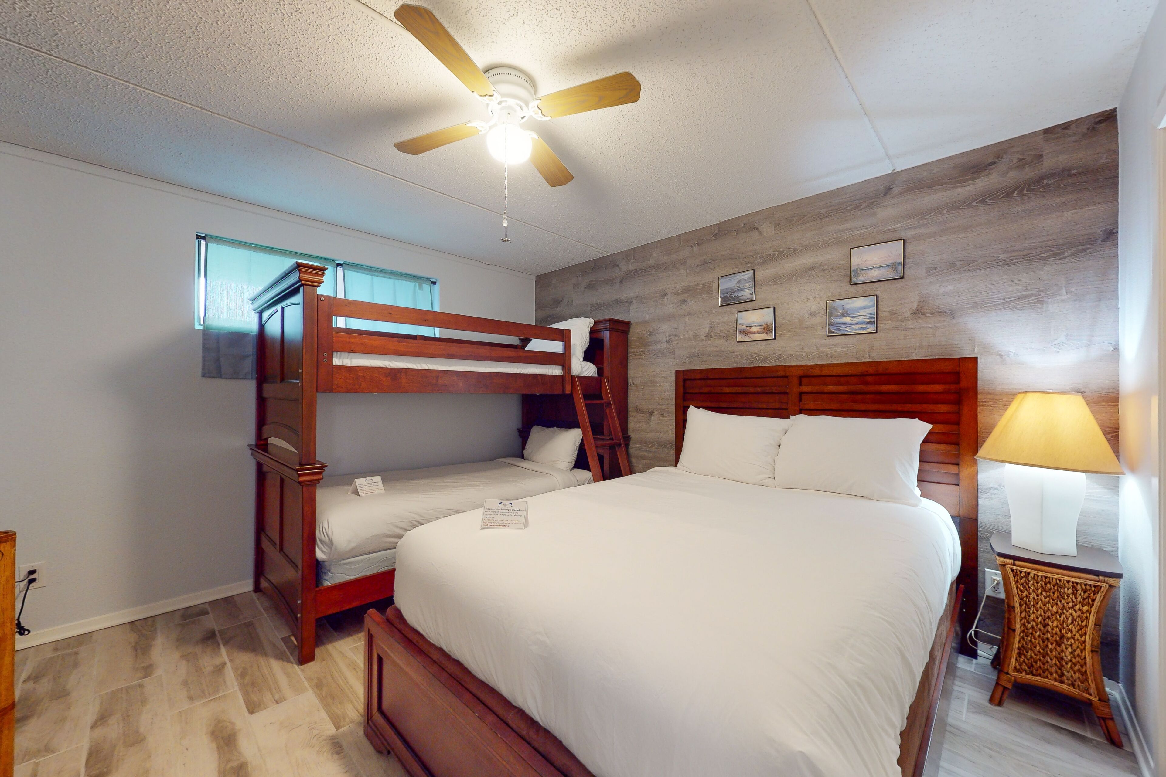 A bedroom with a bunk bed, a single bed, a ceiling fan, a nightstand with a lamp, and a window with blinds.