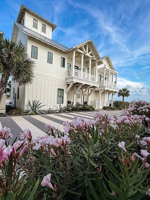 A large, multi-story house with a balcony, surrounded by a garden with pink flowers.