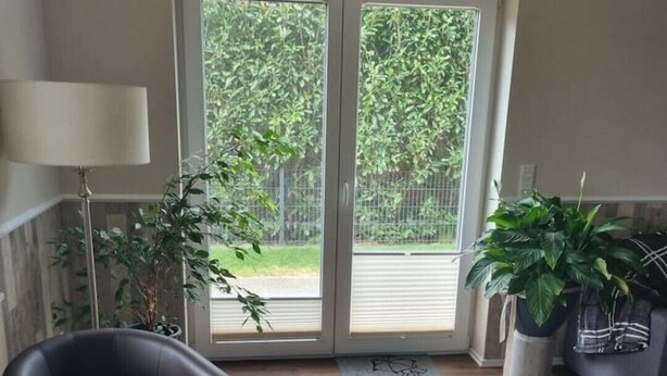 Une pièce dotée d’une grande porte-fenêtre coulissante donnant sur un jardin, d’un fauteuil en cuir noir, d’une petite table ronde ornée d’une plante et d’un tapis en peau de vache sur le sol.
