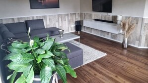 Un salon moderne avec un canapé gris, un téléviseur à écran plat fixé au mur, une console blanche et une grande plante verte.