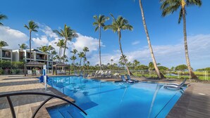 A swimming pool with lounge chairs, palm trees, and a modern building in the background. - Mauna Lani Condo | Short Walk to Beach (Kamuela)