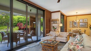A spacious living area with a glass sliding door leading to a garden, a comfortable sofa set, a coffee table, and a dining area with wicker furniture. - Mauna Lani Condo | Short Walk to Beach (Kamuela)