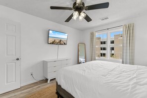 A bedroom with a bed, a ceiling fan, a TV, a dresser, a window with curtains, and a mirror.