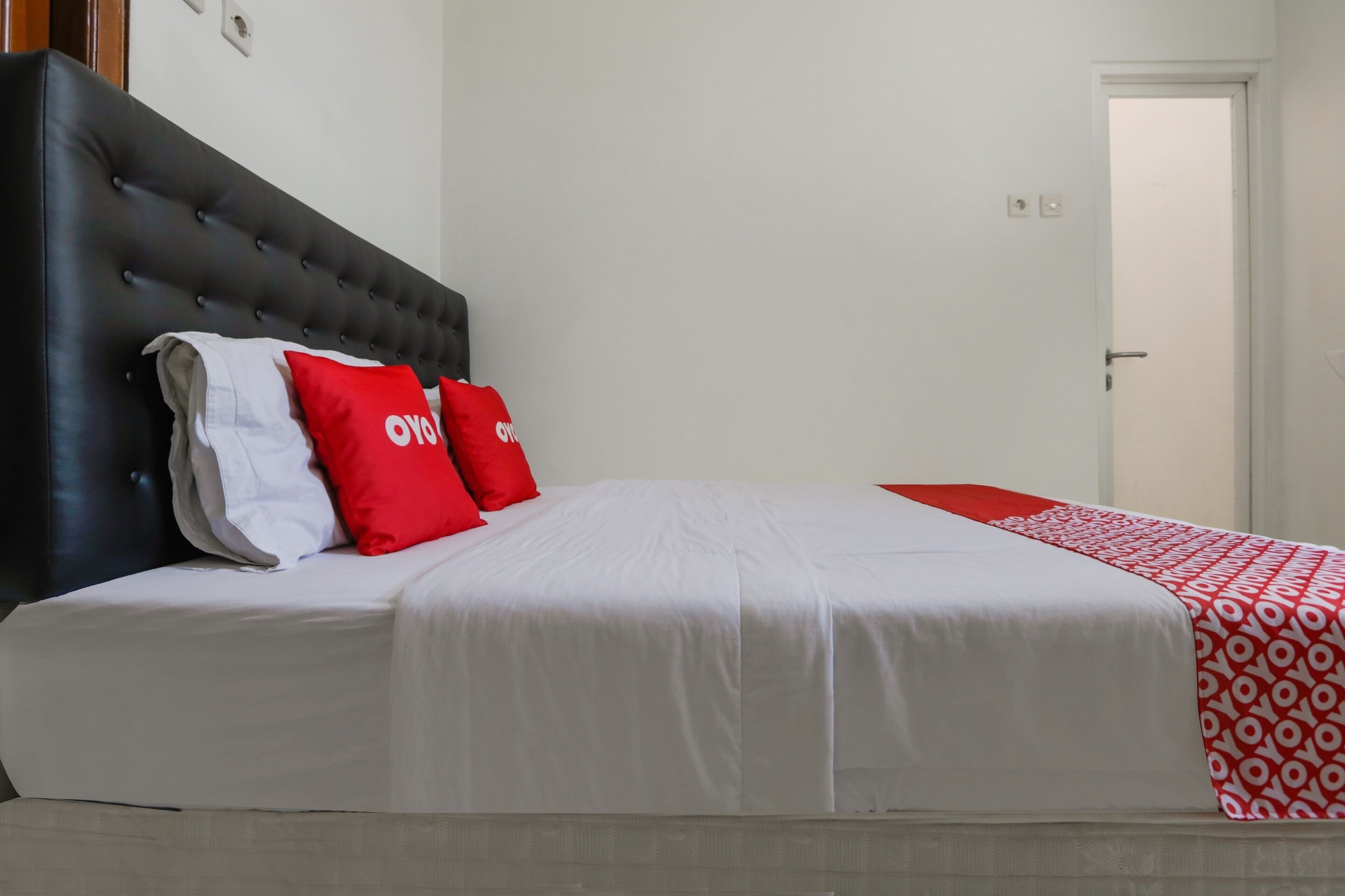 Habitació d'hotel amb un capçal de llit tapissat de color negre, roba de llit blanca i almoines vermelles amb el text "OVYO".