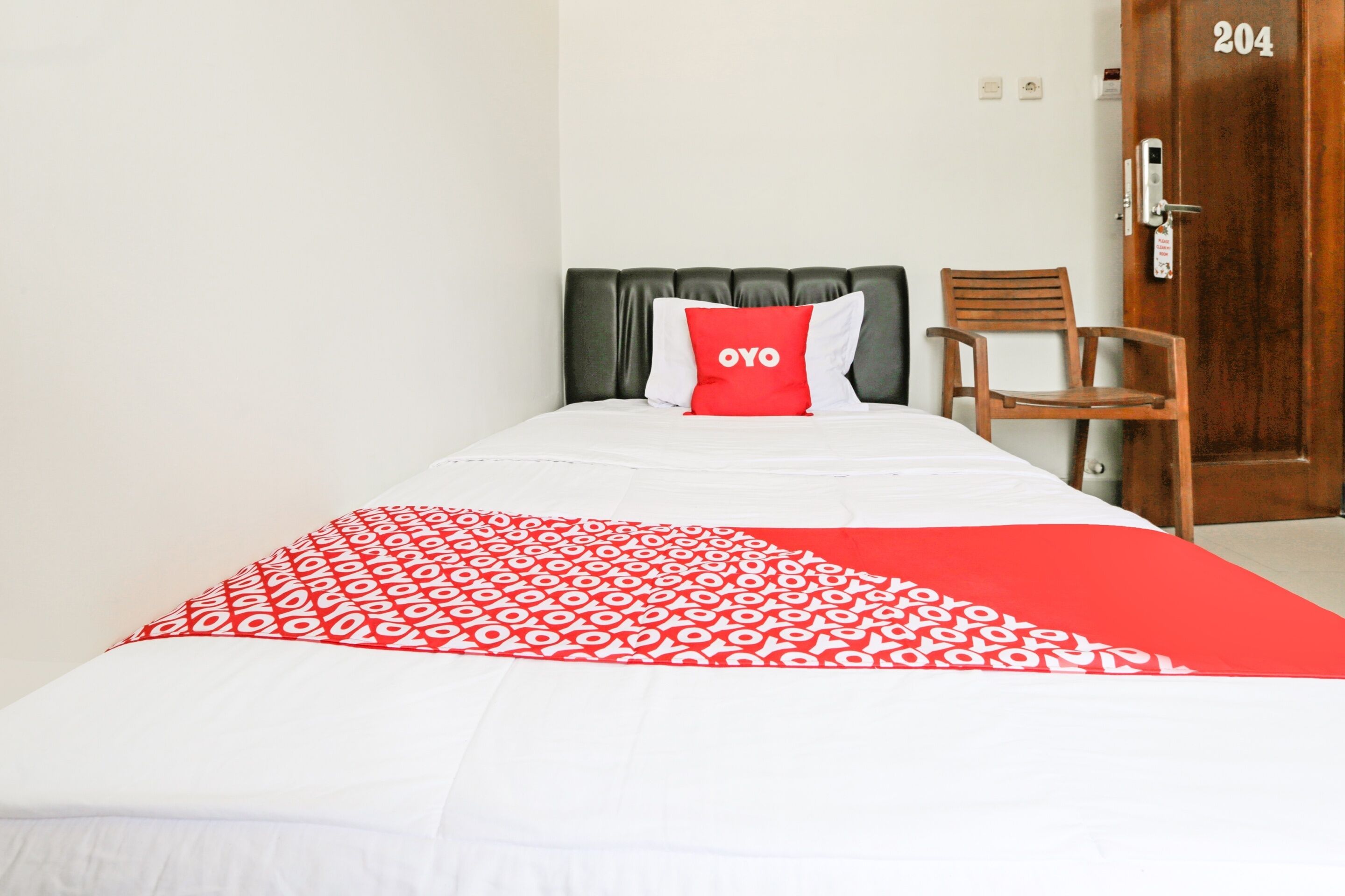 Habitación de hotel con una cama, una colcha con estampado rojo y blanco, un almohadón rojo con el texto "OYO", una silla de madera y una puerta con el número 204.