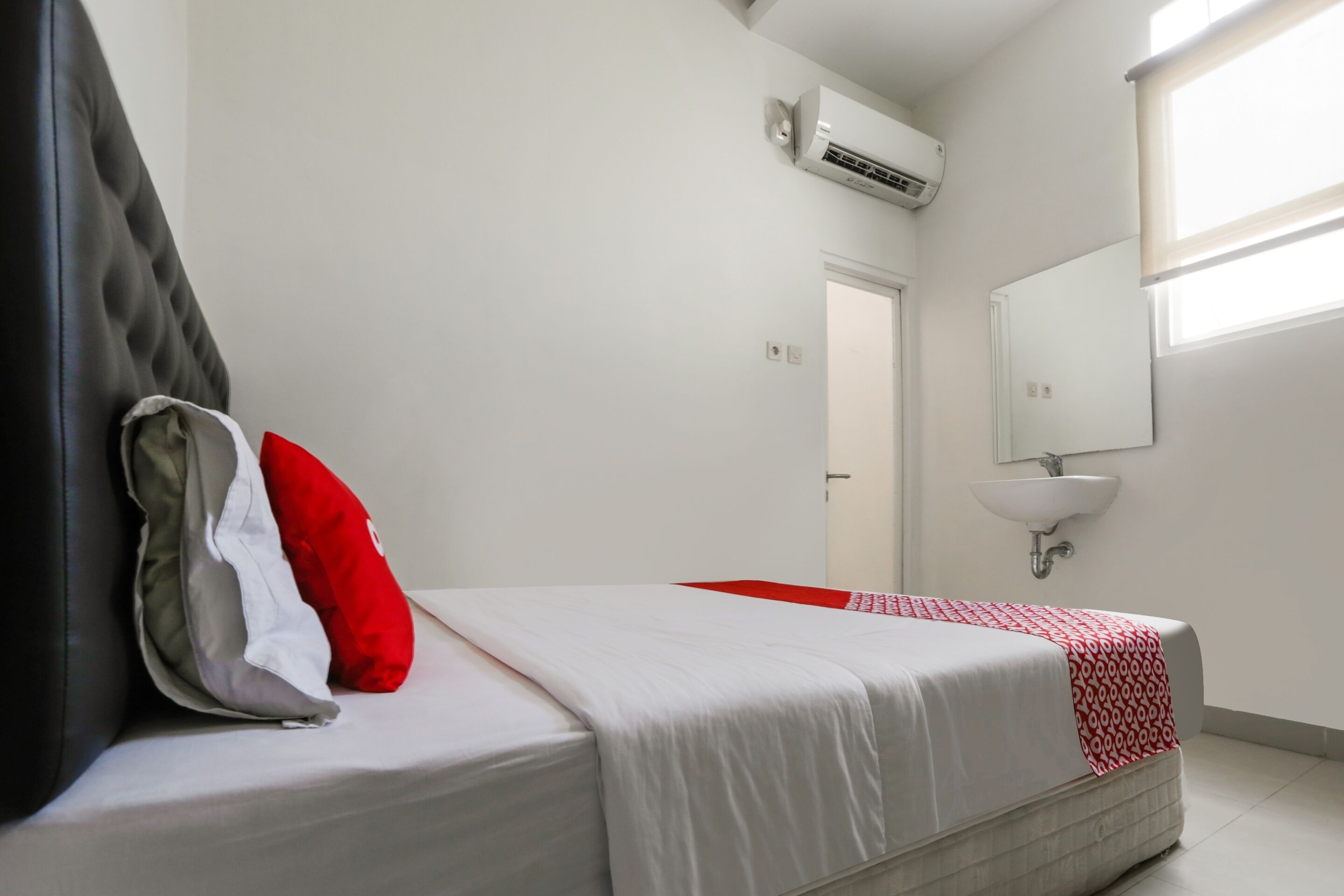 Ein ordentlich bezogenes Bett mit einer rot-weißen Tagesdecke, einem grau-weißen Kopfkissen und einem schwarzen Kopfteil. An der Wand über dem Bett ist eine weiße Klimaanlage montiert, und darüber hängt ein weißes Waschbecken mit Spiegel.