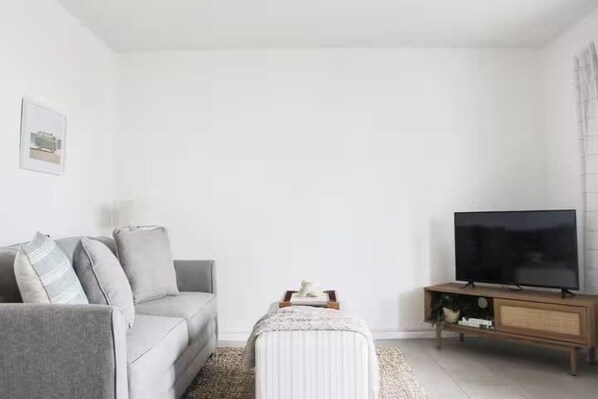 A living room with a grey sofa, a television on a wooden stand, and a small table with a lamp.