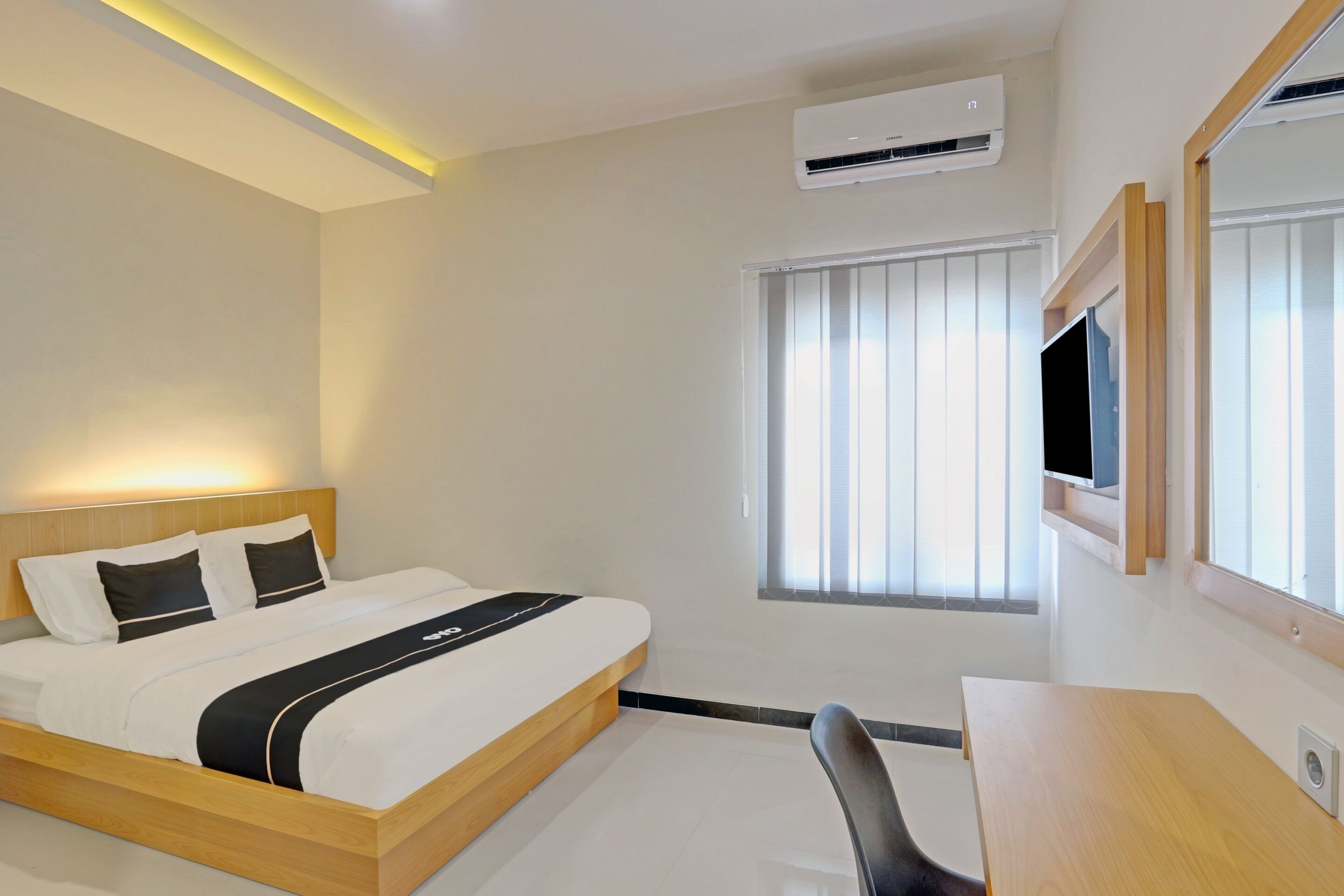 A hotel room with a bed, desk, chair, air conditioning unit, window with blinds, and a mounted TV.