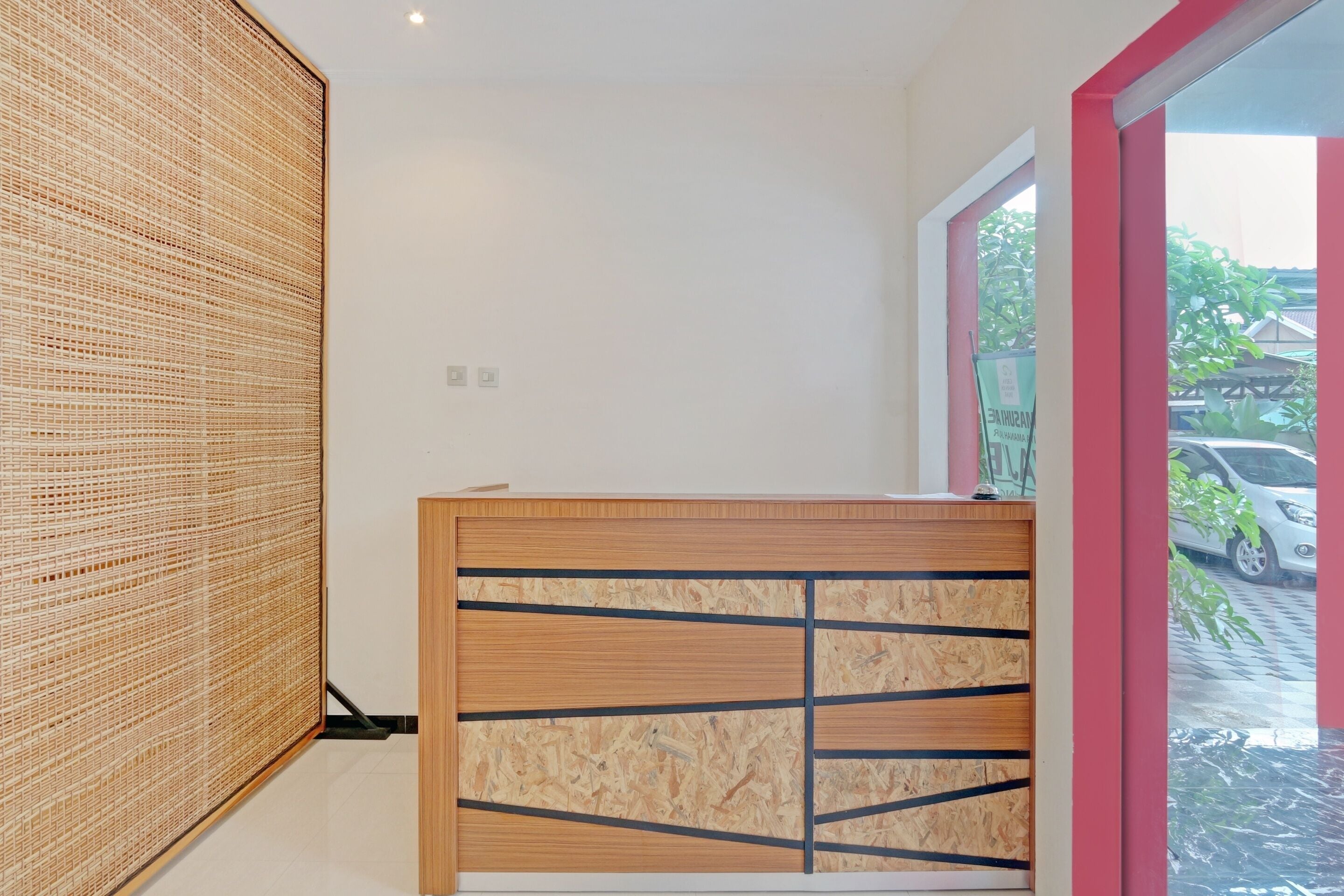 A reception desk with a wooden finish and black horizontal lines, a woven bamboo partition, and a view of parked cars through a glass door.