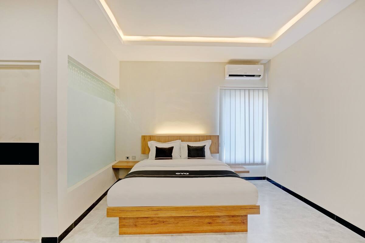 A modern hotel room with a large bed, a bedside table, an air conditioning unit, and a window with blinds.
