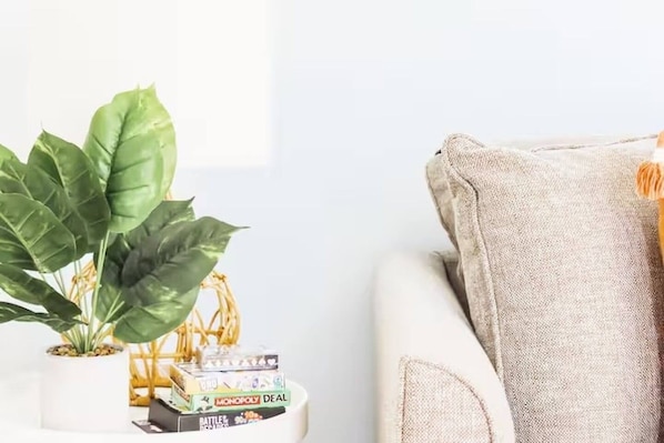 A white sofa with a grey cushion, a small white table with a plant and board games, and a plain wall in the background.