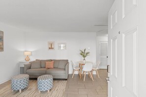 A modern living room with a grey sofa, two ottomans, a small table with chairs, and a lamp. - Central Location l Steps from Ocean l Top Floor (Myrtle Beach)