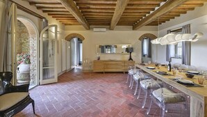 A spacious dining area with a long wooden table, clear chairs, and a rustic brick floor.