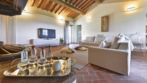 A spacious living room with a high ceiling, a flat-screen TV mounted on the wall, a white sofa, and a tray with wine glasses and bottles.
