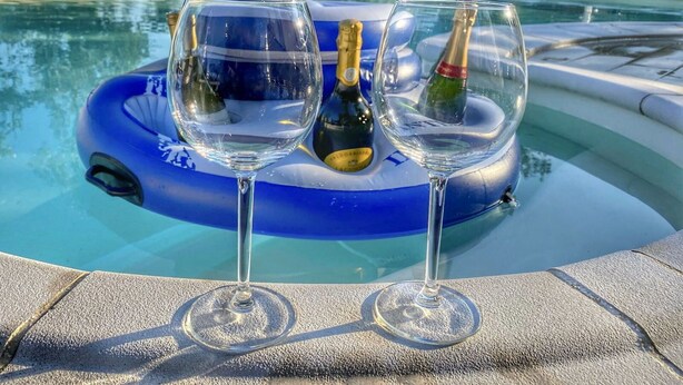 Two wine glasses and a bottle on a blue float in a pool, surrounded by trees and chairs.