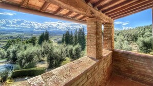 A rustic stone balcony with a wooden roof, overlooking a mountainous landscape with trees and a clear sky.