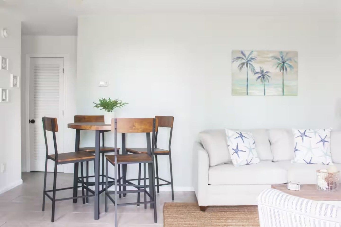 A dining area with a table and chairs, a white sofa with star-patterned cushions, and a painting of palm trees on the wall.