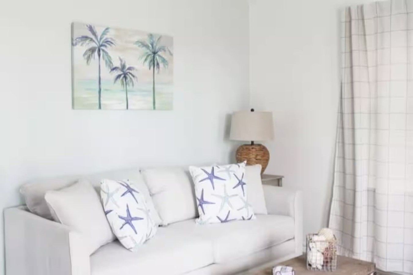 A white sofa with star-patterned cushions, a lamp on a side table, and a painting of palm trees on the wall.