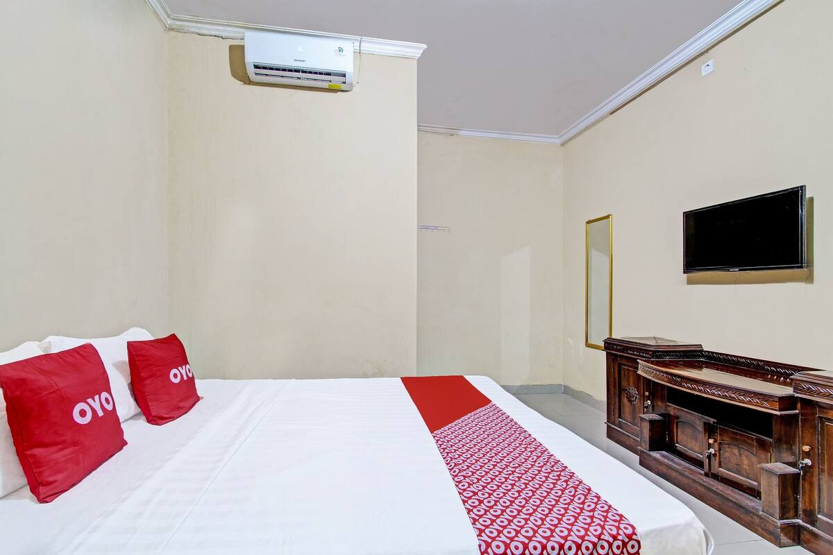 Habitación de hotel con cama, ropa de cama roja y blanca, unidad de aire acondicionado, mueble de madera y televisor de pantalla plana montado en la pared.