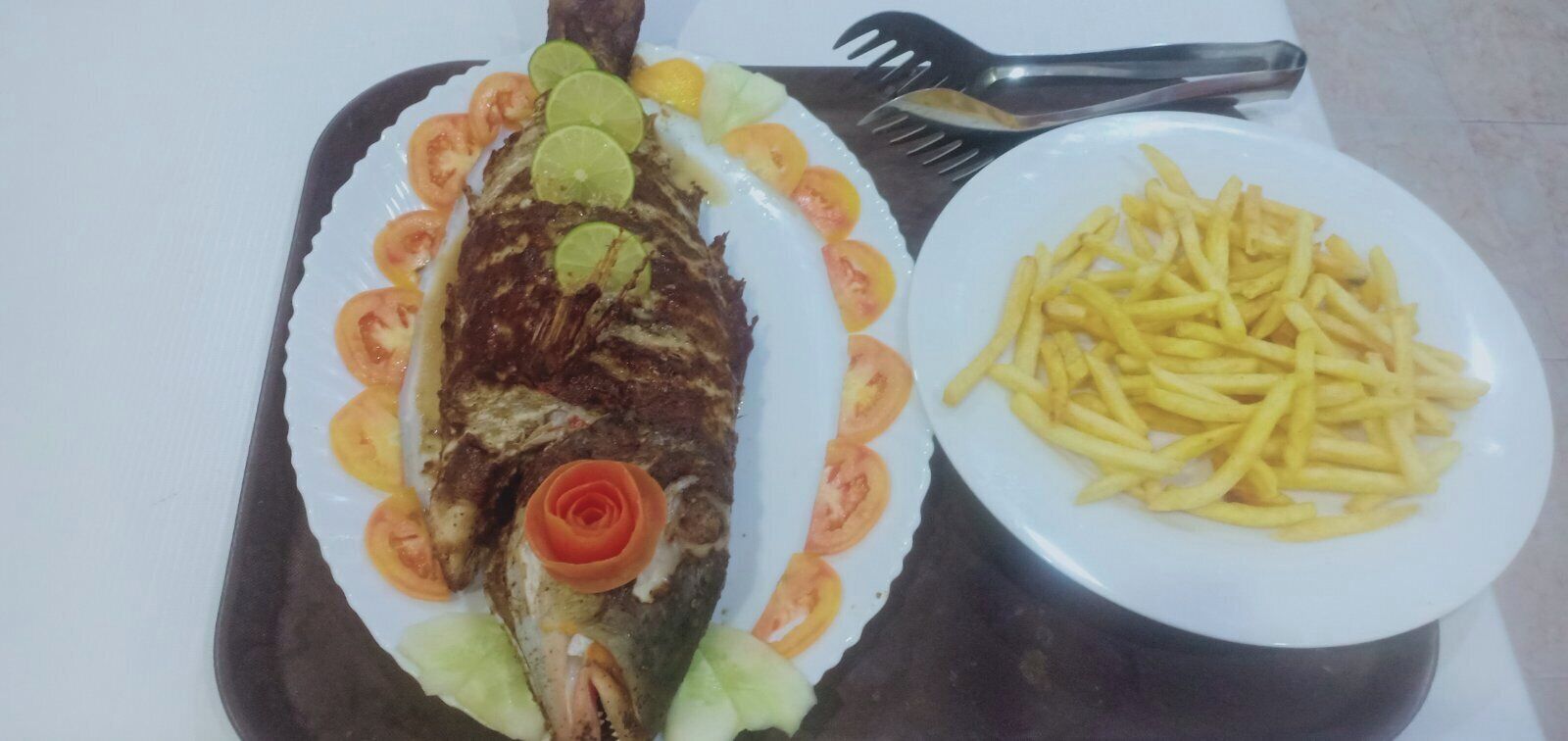 A plate with a whole fish garnished with lime slices and tomato rings, and a separate plate of French fries.