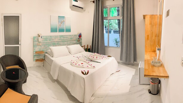 A bedroom with a bed, a chair, a small table, a window with curtains, and a wall-mounted air conditioner. - FINS DIVE AND LEISURE HOTEL (Rasdhoo)