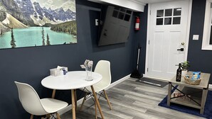 Une pièce avec une table et des chaises blanches, un tableau représentant un paysage de montagne, un extincteur et une porte avec une fenêtre.