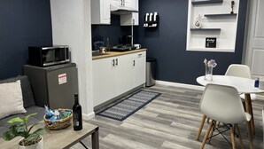 Une cuisine compacte avec des meubles blancs, un four à micro-ondes et un petit réfrigérateur. On y trouve un coin repas avec une table et des chaises blanches, une plante en pot et un vase décoratif.