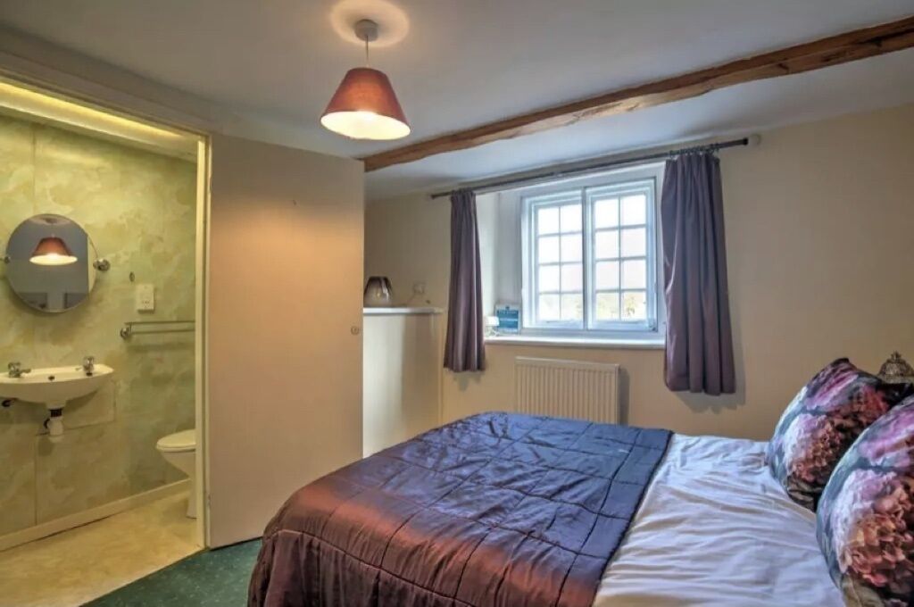 A bedroom with a bed, a window with curtains, a radiator, and a bathroom with a sink and mirror.