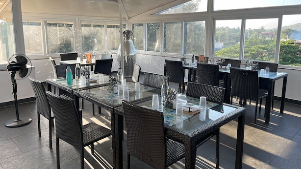 An outdoor dining area with wicker tables and chairs, large windows, and a view of greenery. - Candolim Suites (Candolim)