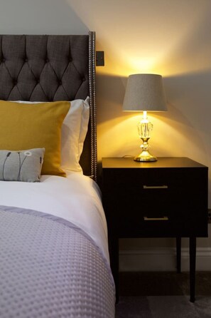 A neatly made bed with a tufted headboard, a bedside table with a lamp, and a small drawer unit.