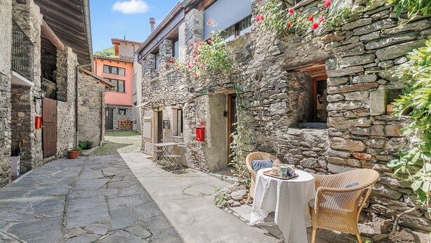A narrow stone pathway leading to a rustic stone building with a small outdoor seating area featuring a wicker chair and a table. - Rustico Stock With Free Wifi (Monteceneri)