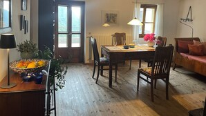 A dining area with a wooden table, chairs, and a vase of flowers. There is a window with curtains and a door leading outside. - Gemütliches Haus mit Blick zum Meer (Glori)