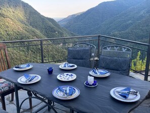 Mesa posta para seis pessoas, com pratos, talheres e uma vela, com vista para uma paisagem montanhosa.