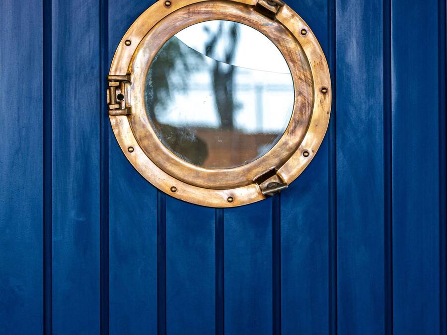 Une petite fenêtre circulaire en laiton, semblable à un hublot de bateau, avec un loquet articulé, est fixée sur une porte bleue.