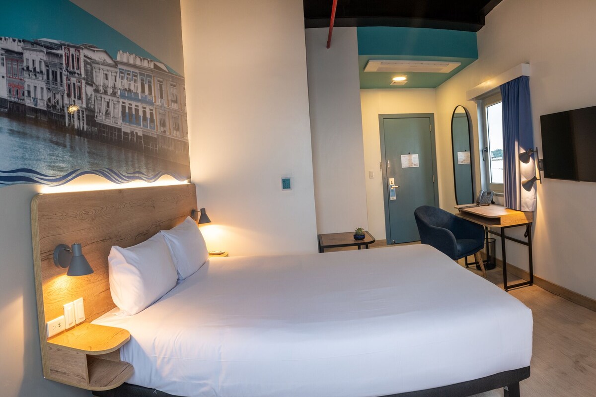 Habitación de hotel con cama, mesita de noche, lámpara, escritorio con silla y televisor montado en la pared.