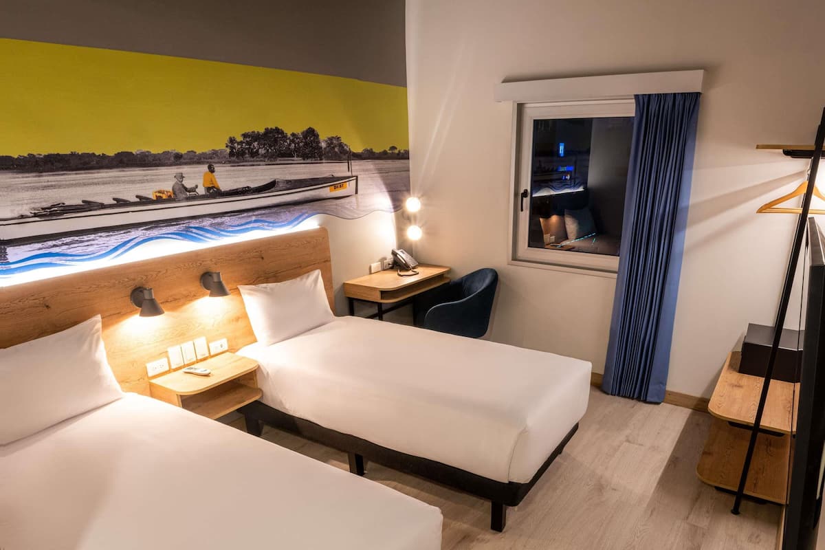 Habitació d'hotel amb dos llits, una taula de treball, una cadira, una làmpada de taula, una nitzeta i una finestra amb cortines, a més d'un mural a la paret.