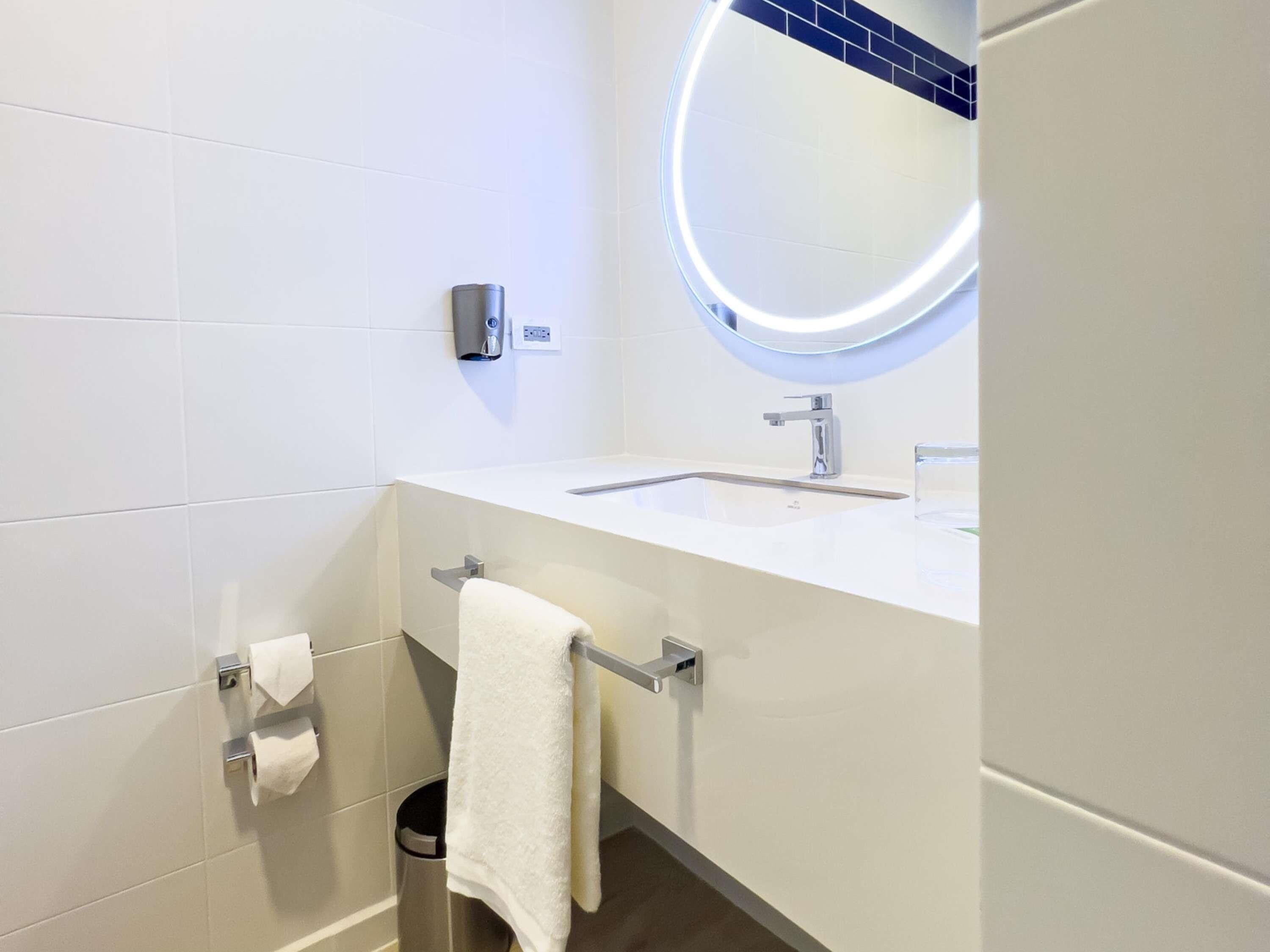 A modern bathroom with a white sink, a round mirror with under-cabinet lighting, a towel rack, and a paper towel dispenser.