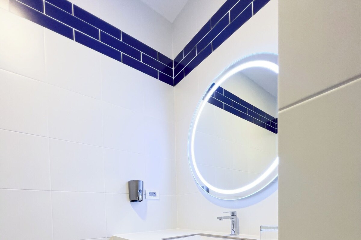 Un baño moderno con un lavamanos blanco, un espejo con luz circular, un perchero y un cubo de basura.