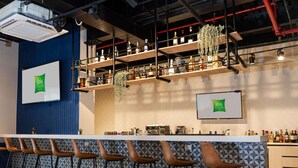 A bar area with a long counter, multiple stools, and shelves with bottles. - Ibis Styles El Malecon Guayaquil (Guayaquil)