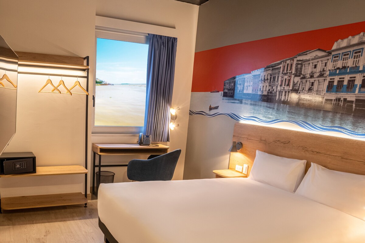 Habitación de hotel con una cama grande, un escritorio con teléfono, una silla, una mesita con planta y una ventana con vista a un cuerpo de agua y edificios.