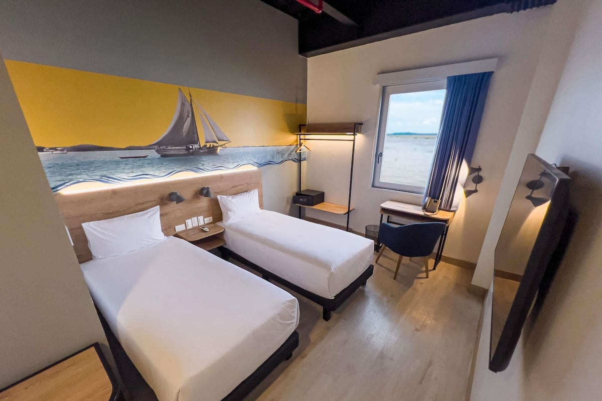 Habitació d'hotel amb dos llits, una taula de treball, una cadira, una finestra amb vista al mar i un mural d'un veler.