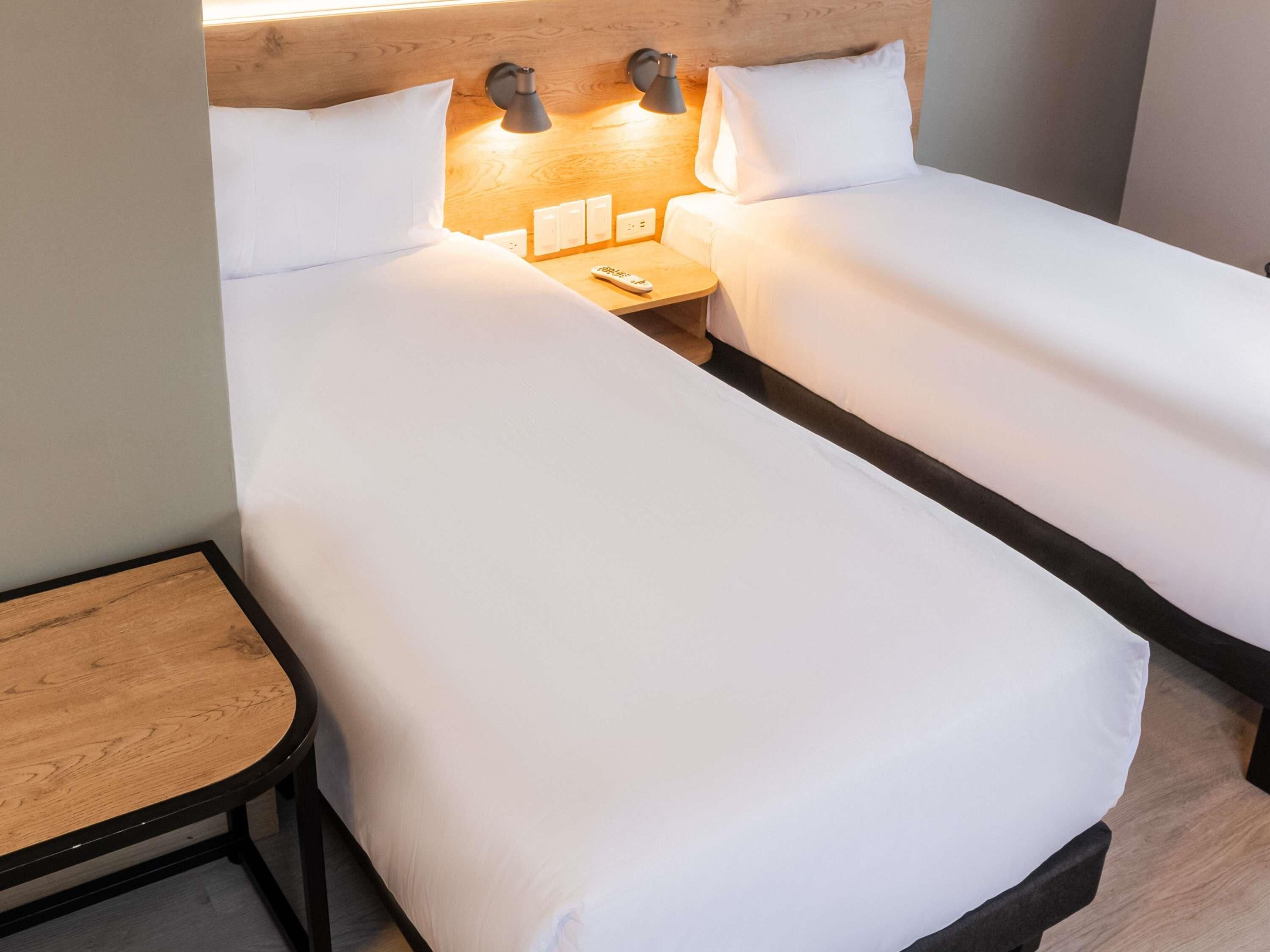 Quarto de hotel com duas camas de solteiro, uma pequena mesa de madeira e uma mesinha de cabeceira com luminária e controle remoto.