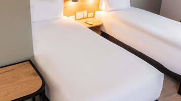 Quarto de hotel com duas camas de solteiro, uma pequena mesa de madeira e uma mesinha de cabeceira com luminária e controle remoto.