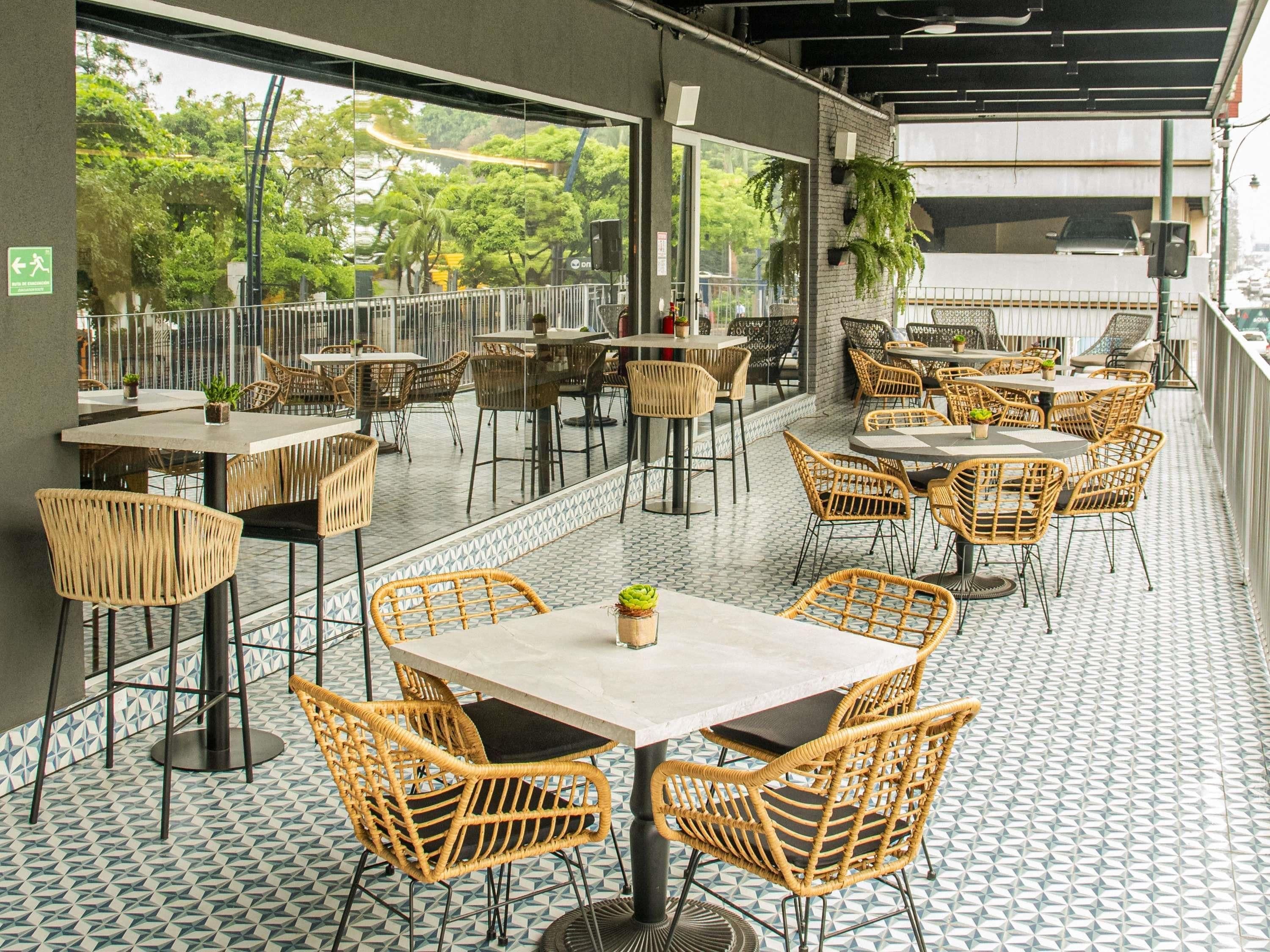 Un espace de restauration en plein air avec des chaises et des tables en osier, un sol carrelé et une vue sur la verdure grâce à de grandes fenêtres.