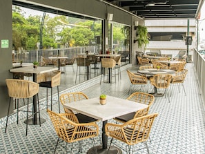 Un espace de restauration en plein air avec des chaises et des tables en osier, un sol carrelé et une vue sur la verdure grâce à de grandes fenêtres.